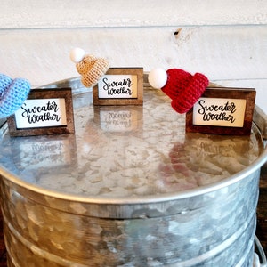Puede incluir: Tres marcos de madera peque&ntilde;os con el texto "Sweater Weather" en cada uno. Cada marco tiene un peque&ntilde;o gorro de punto de un color diferente: azul, tostado y rojo.