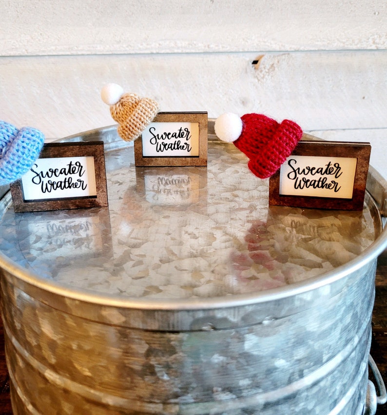 Puede incluir: Tres marcos de madera peque&ntilde;os con el texto "Sweater Weather" en cada uno. Cada marco tiene un peque&ntilde;o gorro de punto de un color diferente: azul, tostado y rojo.