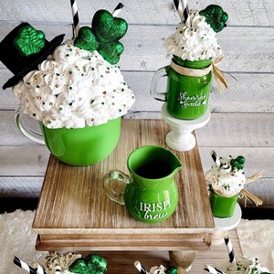 May include: A set of five green and white ceramic mugs with whipped cream and shamrock decorations. The mugs are arranged on a tiered wooden stand. The mugs are decorated with the text "Shamrock Sweetie" and "Irish Brew".
