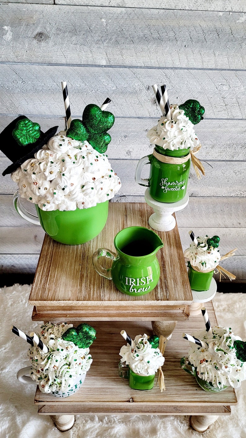 May include: A set of five green and white ceramic mugs with whipped cream and shamrock decorations. The mugs are arranged on a tiered wooden stand. The mugs are decorated with the text "Shamrock Sweetie" and "Irish Brew".