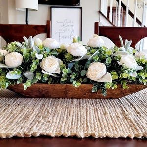 May include: A wooden dough bowl filled with a floral arrangement of white flowers and green foliage. The bowl is sitting on a woven table runner.