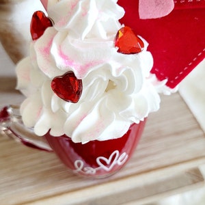 May include: A red mug with white lettering that says "love" and a heart. The mug is filled with white whipped cream and topped with red heart-shaped embellishments. A red felt envelope with a pink heart is on top of the whipped cream.
