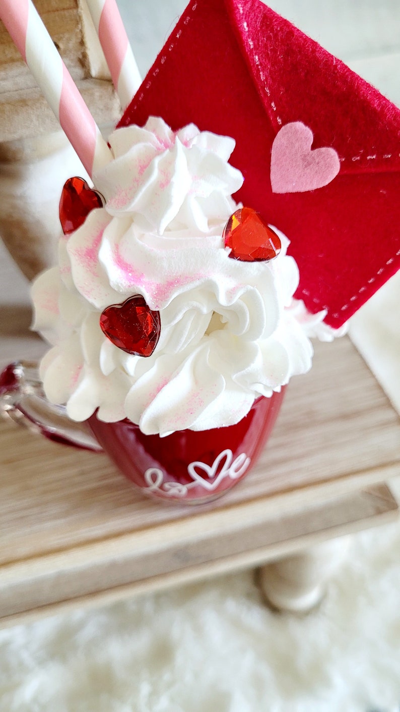 May include: A red mug with white lettering that says "love" and a heart. The mug is filled with white whipped cream and topped with red heart-shaped embellishments. A red felt envelope with a pink heart is on top of the whipped cream.
