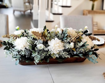 May include: A long, rectangular wooden dough bowl filled with an arrangement of white peonies, small white ranunculus, green eucalyptus leaves, and textured golden-brown floral sprigs. The centerpiece features a dense mix of foliage.