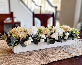 May include: A rectangular white wooden box overflowing with a floral arrangement. The arrangement features cream, white, and light green flowers, with dark green foliage. The centerpiece sits on a woven beige placemat.