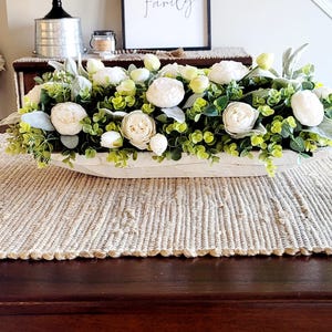 May include: White wooden dough bowl filled with a floral arrangement of white flowers and green foliage. The bowl is sitting on a woven beige table runner on a dark wood table. A sign with the text "Some call it chaos we call it family" is hanging on the wall behind the bowl.