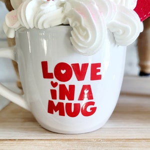 May include: White ceramic mug with red lettering that says "LOVE in a MUG" with a small red heart in each letter. The mug is filled with white whipped cream and a red candy.