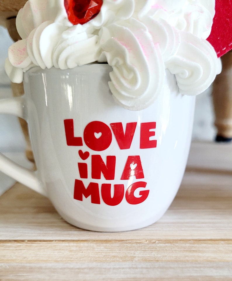 May include: White ceramic mug with red lettering that says "LOVE in a MUG" with a small red heart in each letter. The mug is filled with white whipped cream and a red candy.