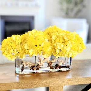 May include: A rectangular clear glass vase containing bright yellow hydrangea blossoms and natural driftwood pieces. The vase features a grid-like metal frame structure that holds the flowers and wood in place within the transparent glass container.