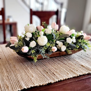 May include: A wooden dough bowl filled with a floral arrangement of white and pink flowers, greenery, and small white berries. The bowl is sitting on a woven brown table runner.