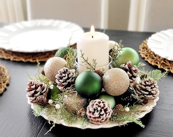 May include: A centerpiece featuring a white pillar candle in a glass holder surrounded by green and gold ornaments, frosted pinecones, and evergreen sprigs on a scalloped white plate. The arrangement includes matte and glittery finishes.