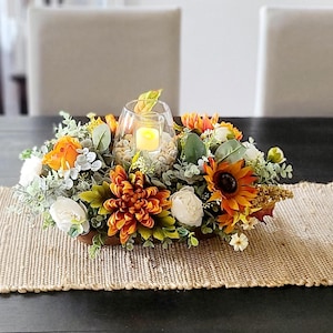 May include: A decorative centrepiece featuring a wooden bowl filled with an arrangement of orange, yellow, and white artificial flowers, including sunflowers and chrysanthemums. A glass candle holder with a lit candle sits in the centre, surrounded by small stones. The arrangement rests on a woven table runner.