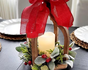 May include: A wooden lantern frame featuring a large red burlap bow at the top, containing a cream-colored pillar candle surrounded by a base of green foliage, silver-toned leaves, and small red berries, creating a decorative centerpiece.