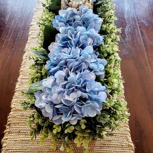 May include: A blue hydrangea centerpiece with greenery on a burlap table runner. The centerpiece includes a glass vase with a candle and rocks.