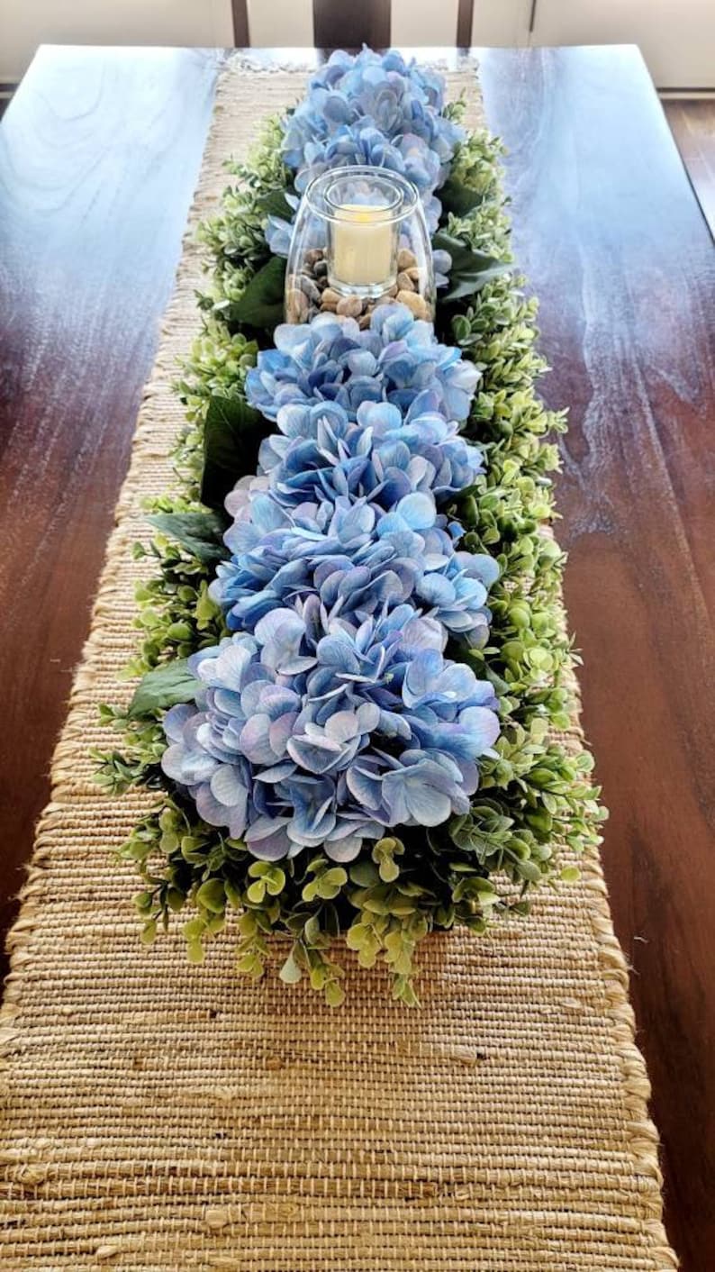 May include: A blue hydrangea centerpiece with greenery on a burlap table runner. The centerpiece includes a glass vase with a candle and rocks.