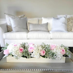 May include: A white wooden box filled with a floral arrangement of pink roses and white hydrangeas. The arrangement is on a white coffee table with a decorative border.