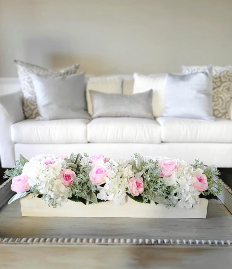 May include: A white wooden box filled with a floral arrangement of pink roses and white hydrangeas. The arrangement is on a white coffee table with a decorative border.