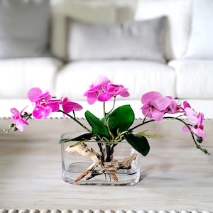 May include: A glass vase with a square base filled with water and a piece of driftwood. The vase is decorated with pink orchid flowers and green leaves.