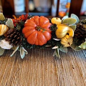 May include: A fall-themed centerpiece with a faux pumpkin, gourds, pinecones, sunflowers, and white flowers. The centerpiece is arranged on a brown woven surface.