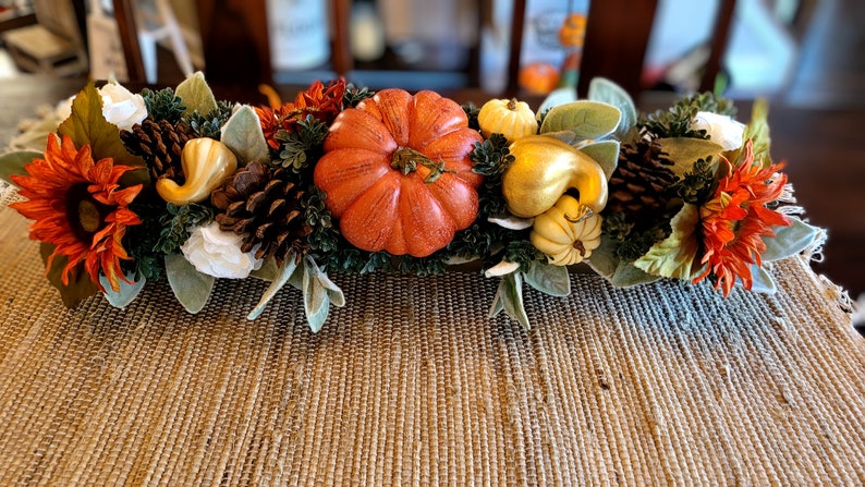 May include: A fall-themed centerpiece with a faux pumpkin, gourds, pinecones, sunflowers, and white flowers. The centerpiece is arranged on a brown woven surface.