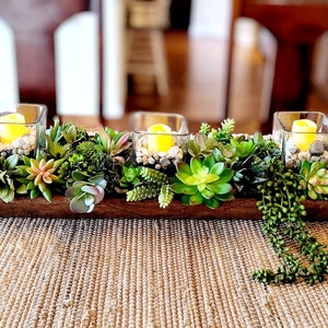 May include: A wooden trough centrepiece filled with artificial succulents and three glass candle holders with lit tea lights.