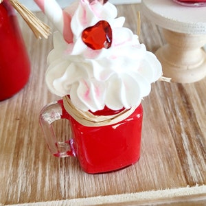 May include: A red glass jar with a clear handle filled with a red liquid topped with white whipped cream and pink and red heart-shaped decorations. The jar is sitting on a wooden surface.