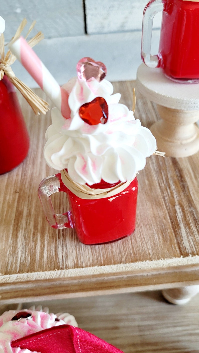 May include: A red glass jar with a clear handle filled with a red liquid topped with white whipped cream and pink and red heart-shaped decorations. The jar is sitting on a wooden surface.
