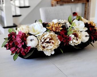 May include: A long black decorative dough bowl filled with an arrangement of artificial flowers and small white pumpkins. The floral display features white magnolias, cream peonies, deep red hydrangeas, and golden-brown autumn blooms.
