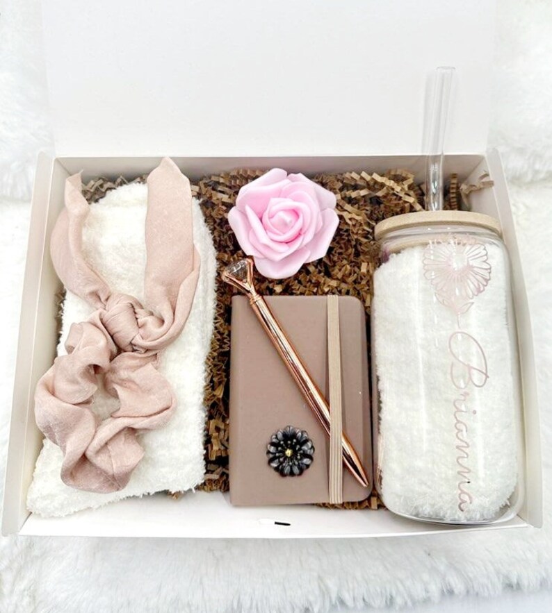 May include: A gift box filled with a white fuzzy headband, a pink rose, a brown notebook with a rose gold pen, and a clear glass tumbler with a straw and the name "Brianna" written on it.