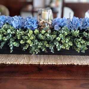 May include: A wooden box centerpiece with blue hydrangea flowers and green foliage. The box is filled with small rocks and a glass candle holder.