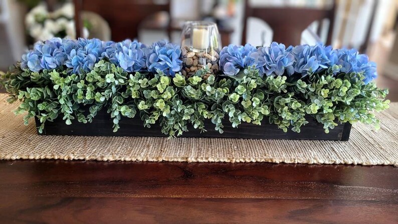 May include: A wooden box centerpiece with blue hydrangea flowers and green foliage. The box is filled with small rocks and a glass candle holder.