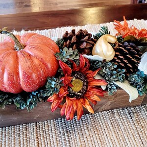 May include: A wooden box filled with fall decor, including a large orange pumpkin, a sunflower, pine cones, and white and gold gourds. The box is sitting on a woven placemat.