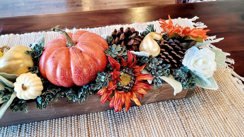 May include: A wooden box filled with fall decor, including a large orange pumpkin, a sunflower, pine cones, and white and gold gourds. The box is sitting on a woven placemat.