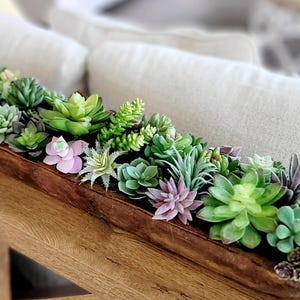 Può includere: Una ciotola in legno marrone scuro, lunga e stretta, riempita con un assortimento di piante succulente artificiali in varie tonalità di verde, rosa chiaro e viola. Le piante sono densamente raggruppate, con texture e forme diverse.