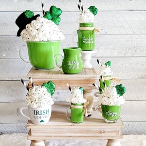 May include: A tiered wooden stand with four green and white mugs with whipped cream and shamrock decorations. The mugs are labeled "Irish Coffee", "Irish Creme", "Shamrock Sweet", and "Lucky".