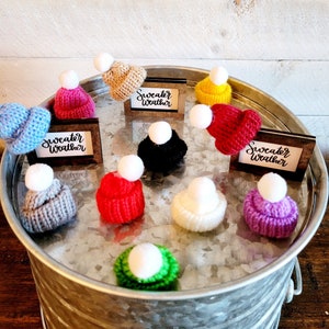 Puede incluir: Una colecci&oacute;n de diez gorros de punto en varios colores, incluyendo rojo, azul, verde, morado, amarillo y blanco, con un pomp&oacute;n blanco en la parte superior de cada gorro. Los gorros est&aacute;n dispuestos en c&iacute;rculo sobre una bandeja plateada con las palabras "Sweater Weather" escritas en tres peque&ntilde;as placas de madera.