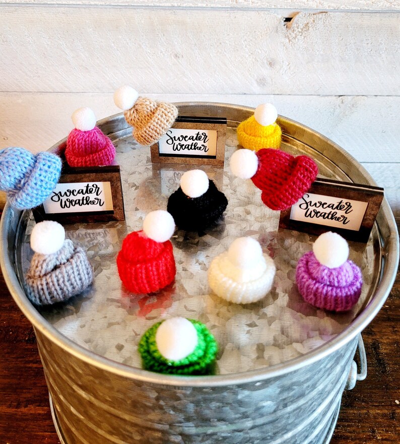 Puede incluir: Una colecci&oacute;n de diez gorros de punto en varios colores, incluyendo rojo, azul, verde, morado, amarillo y blanco, con un pomp&oacute;n blanco en la parte superior de cada gorro. Los gorros est&aacute;n dispuestos en c&iacute;rculo sobre una bandeja plateada con las palabras "Sweater Weather" escritas en tres peque&ntilde;as placas de madera.