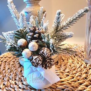 May include: A white glass jar with a white ribbon and the text "Herta Glass" on the front. The jar is filled with a small arrangement of pine sprigs, pine cones, and gold-colored berries. The arrangement is dusted with white faux snow.