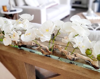 May include: A long, rectangular glass vase containing a series of white orchid stems arranged along a piece of natural driftwood. The white orchid flowers feature yellow centers and green buds, positioned horizontally within the clear glass container.
