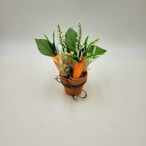 May include: A small terracotta pot with a brown twine bow, filled with faux carrots and green and white foliage.