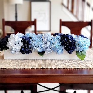 以下が含まれることがあります： 青い花と濃い青色のアジサイの花が植えられた白い長方形のプランター。プランターは、籐製のランチョンマットが敷かれた木製のテーブルの上に置かれています。