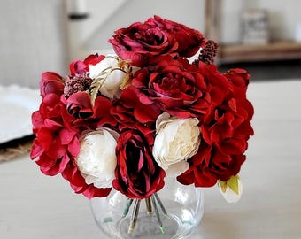May include: A floral arrangement featuring deep red and white roses and peonies in a clear, spherical glass vase. The bouquet includes dark red petals, creamy white blossoms, and small brown textured accents, with visible green stems.