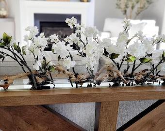 May include: A long, rectangular clear glass vase filled with water, containing several branches of white cherry blossoms and pieces of light brown driftwood arranged horizontally along the bottom of the vessel.