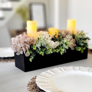 May include: A rectangular black wooden centerpiece box filled with artificial beige hydrangea flowers, white blossoms, and green foliage, accented by three glowing yellow pillar candles. A white scalloped plate sits on a woven placemat nearby.