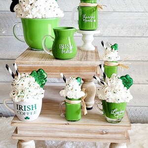 May include: A set of four green and white mugs with shamrock and Irish-themed designs. The mugs are arranged on two wooden tiered stands. The mugs are filled with whipped cream and green liquid, resembling Irish coffee and milkshakes.