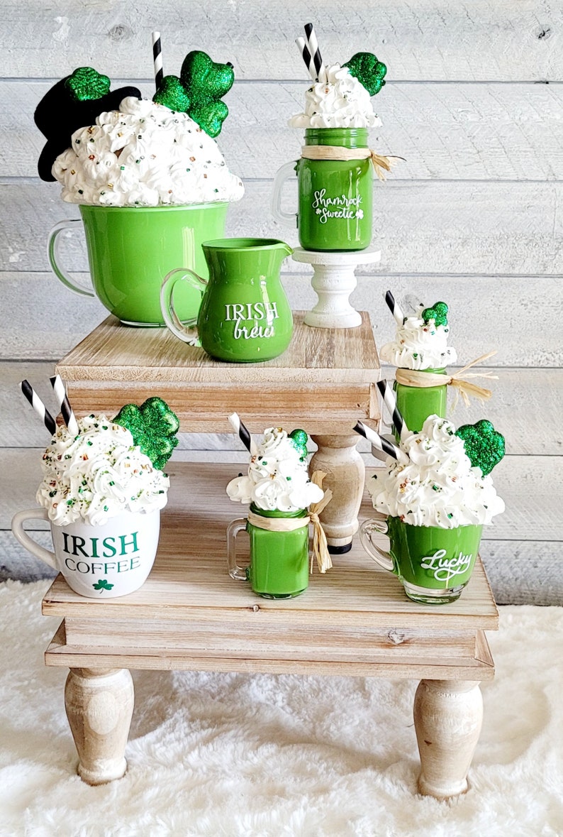 May include: A set of four green and white mugs with shamrock and Irish-themed designs. The mugs are arranged on two wooden tiered stands. The mugs are filled with whipped cream and green liquid, resembling Irish coffee and milkshakes.