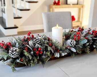 May include: A long wooden dough bowl centerpiece filled with frosted evergreen branches, pinecones, and bright red berries. A single white pillar candle in a glass holder sits in the center of the arrangement, creating a festive display.