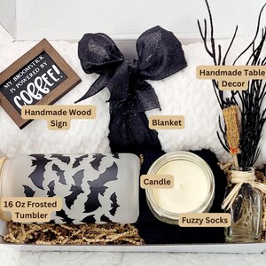 Puede incluir: Una caja de regalo con tem&aacute;tica de Halloween con una manta blanca, un lazo blanco y negro, una vela blanca y negra, un vaso escarchado de 473 ml con un dise&ntilde;o de murci&eacute;lago, un letrero de madera hecho a mano que dice "My broomstick is powered by coffee!" y una peque&ntilde;a botella de vidrio con una decoraci&oacute;n de rama negra.