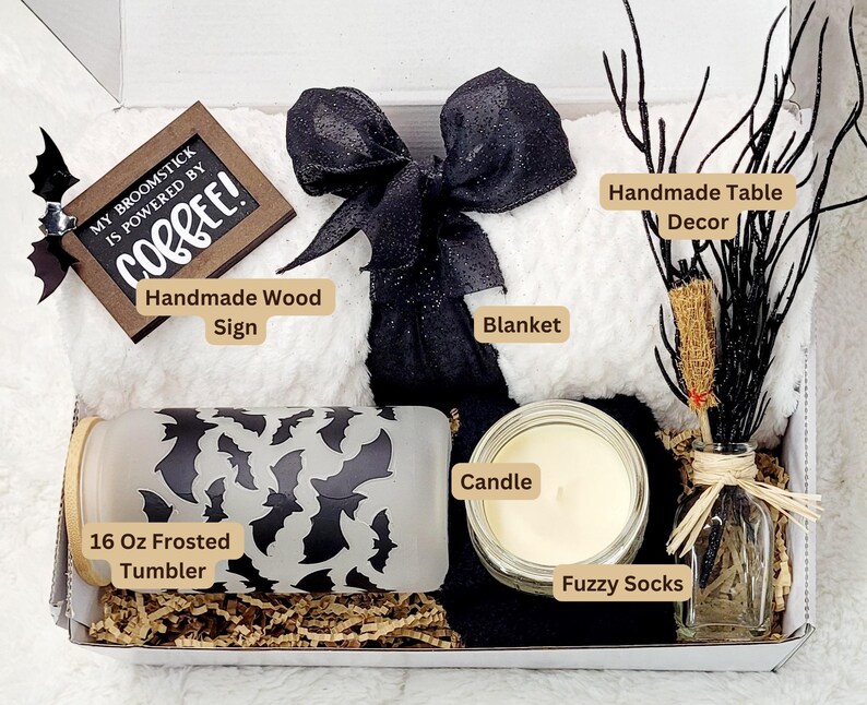 Puede incluir: Una caja de regalo con tem&aacute;tica de Halloween con una manta blanca, un lazo blanco y negro, una vela blanca y negra, un vaso escarchado de 473 ml con un dise&ntilde;o de murci&eacute;lago, un letrero de madera hecho a mano que dice "My broomstick is powered by coffee!" y una peque&ntilde;a botella de vidrio con una decoraci&oacute;n de rama negra.