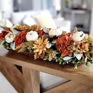 May include: A long, narrow wooden dough bowl filled with an autumn-themed floral arrangement. The centerpiece features white pumpkins, rust-colored hydrangeas, yellow dahlias, green foliage, and small sprigs of dried golden-brown grasses.